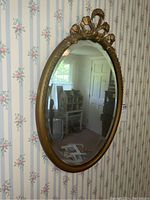 Front view of oval mirror with gold tone frame and decorative ribbon top detail reflecting a floral striped wallpapered room.