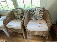 Pair of wicker club chairs with cushions and pillows by Palecek positioned near a window inside a room with hardwood floors.