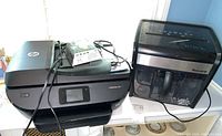 HP ENVY photo printer model 7855 with power cord, HP ink cartridges box, and manuals on top; Staples MailMate shredder beside printer on white table.