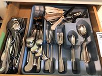 Drawer partial view holding stainless steel flatware including spoons, forks, knives with some wooden handles, arranged in tray and box.