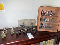 Overview of vintage figurines on dark wood surface including Asian elder figurines, miniature cannons, and clear display box mounted figurines.