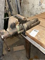 Front-side view of metal bench vise showing rust on handle and worn jaws, clamped to a wooden workbench.