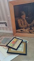 Photo of large framed print of elderly man reading with three smaller framed items and paper underneath on floor