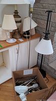 Photo showing group of floor and table lamps on cabinet and floor beside box of lamp parts.