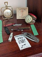 Several pocket cutter tools, a small wristwatch, a vintage travel alarm clock with red foldable cover, and a brass thermometer on a wooden surface.