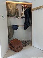 Photo showing closet with multiple olive green army canvas duffle and garment bags hanging, a blue tote bag, and two brown vintage suitcases stacked on the floor.