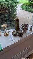 Photo showing seven metal decor items on a windowsill including two vases, an hourglass, two ashtrays, a small box, and a bell.