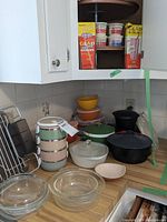 Kitchen corner counter with various bowls and containers including pastel Tupperware nesting bowls, Royal Norfolk Pottery stacking bowls, Pampered Chef micro-cookers, and boxed Glad straws on upper shelf.