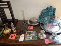 Two silver candlesticks centered in front, silver overlay bowl and covered vegetable bowl with lid on silver trays to the right back, assorted liquor labels and small metal items to the left front, teal cloth napkins behind the bowls and a tall glass vase behind the silver candlesticks.