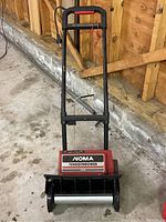 Front view of red and black Noma Turbo Thrower 1500 electric snow thrower showing handle bar, power cord and auger.