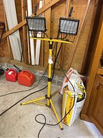 Front view of the yellow twin halogen floodlight with adjustable tripod stand placed inside a garage with some bags and red fuel containers in the background.