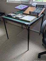 Side and angled view of vintage folding card table showing green playing surface and black folding legs, with various frames and items on top not included in sale.