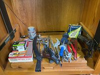 Assorted hand tools arranged on a wooden surface in a storage cabinet. Items include hammer, saw, wrenches, scissors, meter vise, and a box of poultry net staples.