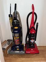 Two upright vacuum cleaners side by side against a wall, one blue and black Eureka Boss 5848 and one red Hoover Feather Light.