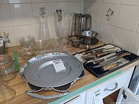 Vintage cutlery and carving utensils with carved handles arranged on trays, metal platters and bowls, glass pitcher, and chafing stand visible