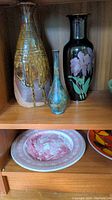 Three ceramic vases on wooden shelf including tall brown beige glazed vase, small blue-green vase, tall black vase with pink floral design.