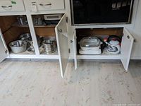 Photo of white cabinets with open cupboard doors showing various pots, pans, electric cooker, and an enamelware rooster tea kettle stored on shelving.