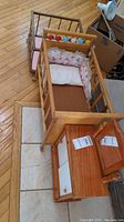 Wooden doll crib with mattress, pillow, and padded lining shown on floor along with dresser and mirror.