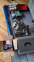 Photo showing film projector, box with reels, blue poles for projection screen, and multiple small boxes and accessories on floor