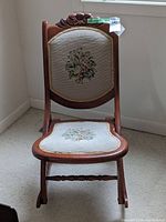 Front view of vintage wood rocking chair showing petit point floral fabric on backrest and seat with carved wood top detail.