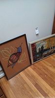 Two framed artworks leaning against the wall on the floor: a colorful abstract bird artwork and a landscape oil painting.