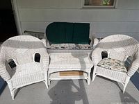 View of the complete set showing two white wicker armchairs with floral seat cushions, loveseat sofa with green and floral cushions, and white wicker coffee table.