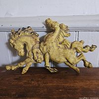 Yellow painted cast iron relief of two horses in a dynamic galloping pose with signs of rust and wear.
