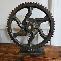 Front view of metal gear sculpture highlighting gear teeth and curved spokes on wooden surface.