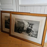 Pair of two framed lithographs leaning against a wall, one showing a fox carrying a goose with a smaller animal, set in oak frames.