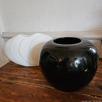 Photo showing glossy black rounded vase and behind it a set of white curved vases arranged in a fan shape on wooden surface.