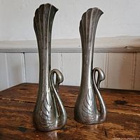 Front view of two silver-plated zinc swan-shaped bud vases showing full profile and height, placed on a wooden table.