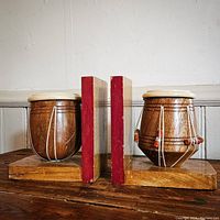 Pair of vintage wooden drum-shaped bookends standing side by side on a wooden surface against a white paneled wall. Both are mounted on rectangular wooden bases with red edges.