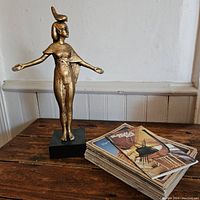 Wide view showing the Egyptian goddess figurine standing on a black base beside a pile of booklets with Egyptian themes on the covers.