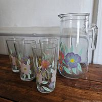 Photo showing four clear glass tumblers with colorful floral designs and one large glass pitcher with matching floral decoration. Set displayed on wooden surface with white background.