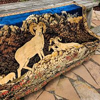 Full view of the rug draped over a bench, showing the overall design with mountain goats and rocky landscape, blue sky background, and floral border.