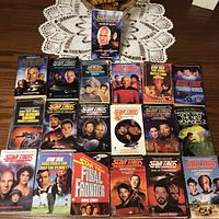 Full lot of 19 Star Trek paperback novels laid out on a table with a white crocheted doily underneath.