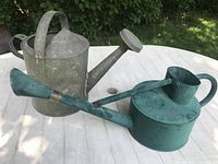 Both metal watering cans placed side by side on a white outdoor table with a background of greenery, showing overall condition and size.