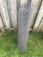 Rolled chainlink fencing leaning against wooden fence on grass.