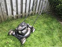 Full side angle showing black Craftsman lawnmower with visible wear on wheels and deck standing on grass with wooden fence background.