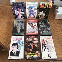 Photo showing 9 Elvis Presley VHS tapes arranged in three rows on a wooden table, showing front covers and boxes in excellent condition.