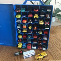 Front view of blue carrying case open showing 48 Hot Wheels cars arranged inside, plus three additional cars in front on wooden table.