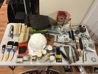 Full view of miscellaneous tools, brushes, hard hat, gloves, and hardware items arranged on a white table for sale