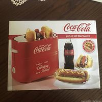 Front of the original box showing red Coca-Cola pop-up hot dog toaster with two hot dogs and buns being toasted.
