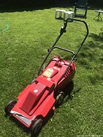 Front and right side view of red electric lawn mower on grass, showing black handle, wheels, and warning label on top.