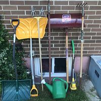 Full view of assorted gardening and lawn tools arranged against a brick wall, including yellow plastic shovel, push broom, cultivators, watering can, and small hand tools