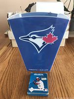 Clear blue Toronto Blue Jays plastic bin with a large Blue Jays bird logo and red maple leaf on front, with package of Joe Carter photos placed at base in front of bin.