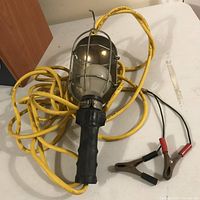 Image showing the entire trouble light with visible metal cage, black handle, long yellow power cord coiled, and battery clamps at the end.