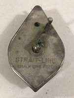 Close-up photo of one side of the diamond-shaped metal chalk line reel, showing the hand crank and embossed brand name