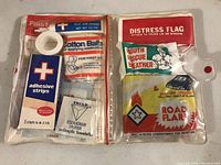Photo showing first aid items including cotton balls, adhesive strips, antiseptic towelettes, distress flag, youth rescue feather, and road flare in original sealed packaging inside a plastic cover with zipper pouch.