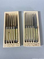 Two sealed wooden boxes each containing six Jean Dubost Laguiole steak knives with cream-colored handles.